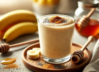 A creamy banana smoothie in a glass, topped with poppy seeds and a dash of cinnamon. The smoothie has a thick texture with a golden hue, placed on a wooden countertop with a banana and honey jar subtly visible in the background.
