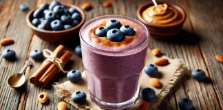 A creamy blueberry smoothie in a glass, blended with almond butter and cinnamon, creating a rich purple hue. The smoothie is garnished with whole blueberries and a sprinkle of cinnamon, set on a rustic wooden surface with almonds and a small dish of almond butter subtly visible in the background.