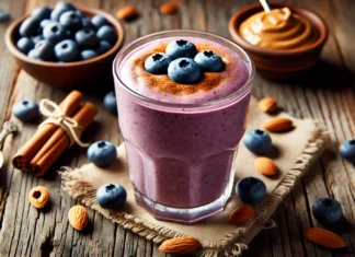 A creamy blueberry smoothie in a glass, blended with almond butter and cinnamon, creating a rich purple hue. The smoothie is garnished with whole blueberries and a sprinkle of cinnamon, set on a rustic wooden surface with almonds and a small dish of almond butter subtly visible in the background.