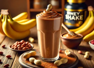 A tall glass of Chocolate-Peanut Power Shake with a rich, creamy brown texture, garnished with a peanut and a light dusting of cocoa powder. Surrounding the smoothie are fresh banana slices, a small bowl of peanut butter, a scoop of chocolate whey protein, and a cinnamon stick on a wooden table, creating a warm and inviting post-workout recovery drink.