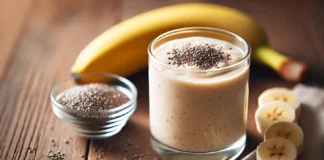 A creamy banana protein smoothie in a clear glass, garnished with chia seeds. A peeled banana and a small bowl of chia seeds sit beside the glass on a wooden table, illuminated by soft natural lighting.