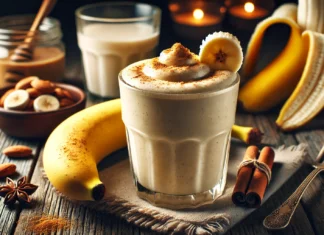 A creamy nutmeg banana smoothie in a clear glass, with a warm and frothy texture, garnished with a sprinkle of nutmeg and a banana slice. The smoothie sits on a rustic wooden table with a halved banana, a small bowl of almond butter, and a glass of warm almond milk, creating a cozy, dimly lit nighttime ambiance.