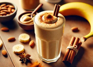 A creamy maca-banana smoothie in a tall glass, garnished with a sprinkle of cinnamon. The smoothie has a smooth, rich texture and a golden hue. Surrounding the glass are a ripe banana, a small bowl of almond butter, and a few cinnamon sticks, all placed on a wooden countertop with soft, warm lighting.