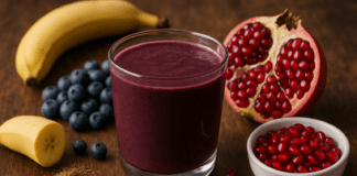 A dark purple acai smoothie surrounded by banana, blueberries, flaxseed, and fresh pomegranate, illustrating a heart-protective antioxidant blend on a wooden surface.