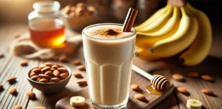 A creamy almond and banana smoothie in a tall glass, garnished with a sprinkle of cinnamon. The smoothie is placed on a wooden countertop with a small bowl of almonds, a bunch of bananas, and a drizzle of honey in the background.