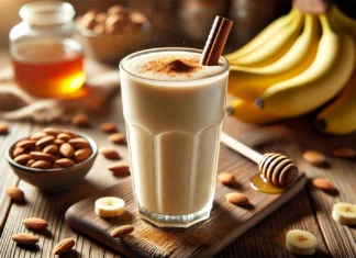 A creamy almond and banana smoothie in a tall glass, garnished with a sprinkle of cinnamon. The smoothie is placed on a wooden countertop with a small bowl of almonds, a bunch of bananas, and a drizzle of honey in the background.