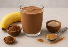 A glass of creamy chocolate smoothie surrounded by ashwagandha powder, cacao, almond butter, and a banana, styled on a warm neutral background with soft lighting.