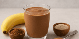 A glass of creamy chocolate smoothie surrounded by ashwagandha powder, cacao, almond butter, and a banana, styled on a warm neutral background with soft lighting.