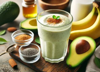 A creamy pale green Avocado Power Fuel Smoothie in a clear glass, surrounded by fresh avocado halves, a ripe banana, chia seeds, honey, cinnamon powder, and almond milk on a wooden surface with soft, natural lighting.