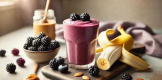A blackberry banana power shake in a glass, featuring a deep purple color and a creamy texture. The smoothie is surrounded by fresh blackberries, a peeled banana, a spoonful of almond butter, and a drizzle of honey. The setting is a bright and modern kitchen with soft natural lighting.