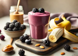 A blackberry banana power shake in a glass, featuring a deep purple color and a creamy texture. The smoothie is surrounded by fresh blackberries, a peeled banana, a spoonful of almond butter, and a drizzle of honey. The setting is a bright and modern kitchen with soft natural lighting.