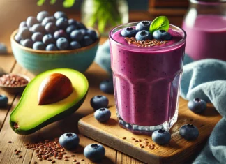 A vibrant blueberry smoothie in a glass, made with blueberries, avocado, flaxseeds, and almond milk, featuring a deep purple color, placed on a wooden surface with fresh blueberries, a halved avocado, and a small bowl of flaxseeds in a softly lit setting.