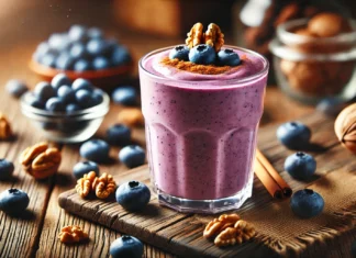 A creamy blueberry smoothie in a clear glass, garnished with walnuts and a sprinkle of cinnamon. The smoothie has a rich purple hue and is placed on a wooden table with fresh blueberries and walnut pieces scattered around, creating a cozy and inviting scene.