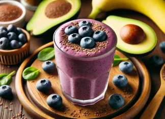 A creamy avocado blueberry smoothie in a clear glass with a rich purple hue, placed on a wooden table. The smoothie is topped with a few fresh blueberries and a sprinkle of flaxseeds. Surrounding the glass are half an avocado, a banana, and a small dish of cinnamon, creating a nutritious and inviting scene.