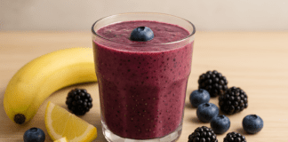 A vibrant berry smoothie in a glass surrounded by banana, lemon wedges, blueberries, blackberries, and chia seeds on a light wooden surface, illustrating a heart-healthy recipe with coconut water.