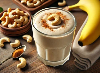 A creamy beige cashew banana smoothie in a clear glass, garnished with a sprinkle of cinnamon and a cashew nut. The background includes a wooden countertop with a banana, a small bowl of raw cashews, a glass of almond milk, and a drizzle of honey.