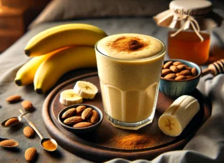 A creamy, golden smoothie in a clear glass, made with fenugreek, banana, almond milk, and cinnamon. The smoothie is garnished with a drizzle of honey and surrounded by fresh banana slices, almonds, a dish of almond butter, and fenugreek seeds in a cozy nighttime setting.