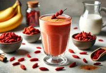 A vibrant red-orange goji berry smoothie in a clear glass, made with banana, almond milk, and cinnamon. Garnished with a few goji berries and cinnamon, set against a kitchen counter with dried goji berries, a banana, a glass of almond milk, and honey.