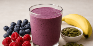 A purple berry smoothie in a glass surrounded by fresh raspberries, blueberries, banana, raw pumpkin seeds, and flaxseed on a neutral surface, highlighting antioxidant-rich and heart-protective ingredients.