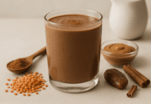 A rich, chocolate-colored lentil smoothie in a clear glass, surrounded by red lentils, almond butter, a date, cinnamon sticks, raw cacao powder, and soy milk on a warm, minimalist background, highlighting its heart-healthy and homocysteine-lowering benefits.