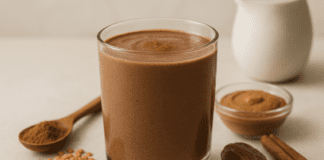 A rich, chocolate-colored lentil smoothie in a clear glass, surrounded by red lentils, almond butter, a date, cinnamon sticks, raw cacao powder, and soy milk on a warm, minimalist background, highlighting its heart-healthy and homocysteine-lowering benefits.