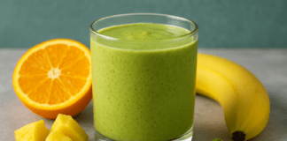 A vibrant matcha citrus smoothie in a glass, surrounded by orange halves, pineapple chunks, a banana, flaxseeds, and a mound of matcha powder on a light gray surface.