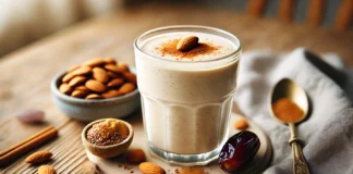 A creamy nutty flax vanilla smoothie in a clear glass, placed on a wooden surface. The smoothie has a smooth, beige texture with a drizzle of almond butter on top and a sprinkle of cinnamon. Whole almonds, a Medjool date, and a small pile of ground flaxseeds are arranged beside the glass. The background is softly blurred, hinting at a cozy kitchen setting.