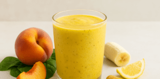 A vibrant golden peach smoothie in a clear glass, surrounded by ripe peach slices, spinach, banana, ginger root, turmeric, and lemon on a clean, calming background, showcasing heart-healthy and energizing ingredients.