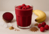 A vibrant red raspberry and beet smoothie in a clear glass, surrounded by fresh raspberries, banana, flaxseeds, ginger powder, ice cubes, and a halved beet on a soft, minimalist background, highlighting heart-healthy ingredients.