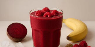 A vibrant red raspberry and beet smoothie in a clear glass, surrounded by fresh raspberries, banana, flaxseeds, ginger powder, ice cubes, and a halved beet on a soft, minimalist background, highlighting heart-healthy ingredients.