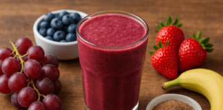 A rich purple-red smoothie in a glass surrounded by fresh red grapes, blueberries, strawberries, banana, and a bowl of ground flaxseed on a wooden surface, highlighting a heart-protective antioxidant blend.
