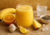 A vibrant yellow smoothie in a clear glass surrounded by banana, orange, lemon wedge, pineapple chunks, garlic, chia seeds, ice cubes, and coconut water on a rustic wooden surface.